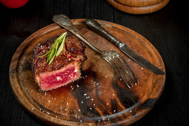 Grill juicy beef steak with salt pepper and rosemary on a wooden board with a knife and fork. Still life