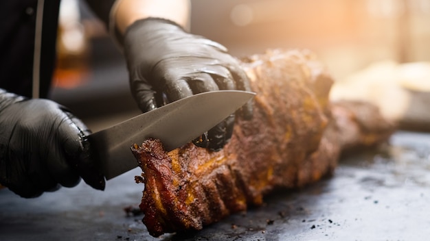 Grill restaurant kitchen. Chef in black cooking gloves using knife to cut smoked pork ribs.