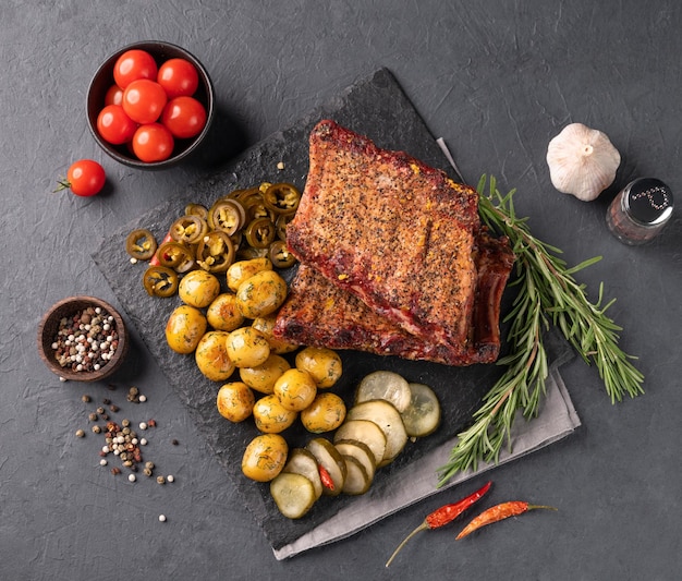 Grilled bbq pork ribs with fried baby potato pickled cucumber and jalapeno and fresh vegetables and herbs on a dark background Concept snacks and food in a bar or restaurant Top view