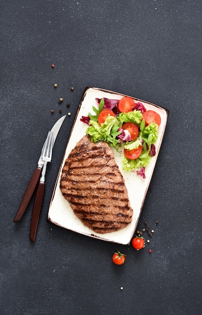 Grilled beef steak in a plate with salad and vegetables on a dark background. Dinner.