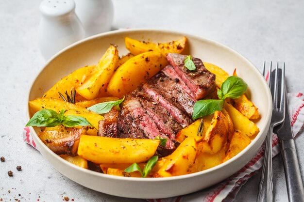 Bistecca di manzo alla griglia con patate e basilico in un piatto bianco su sfondo bianco.