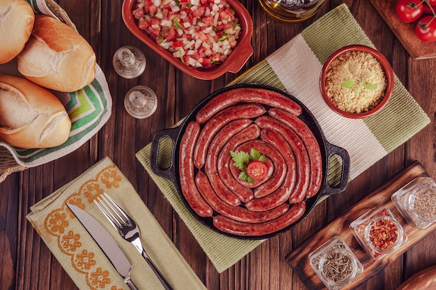 Grilled brazilian sausage on iron plate with bread, salad, farofa and spices