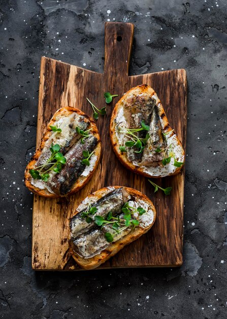 Grilled bread sardines micro greens sandwiches on a rustic\
cutting board on a dark background top view