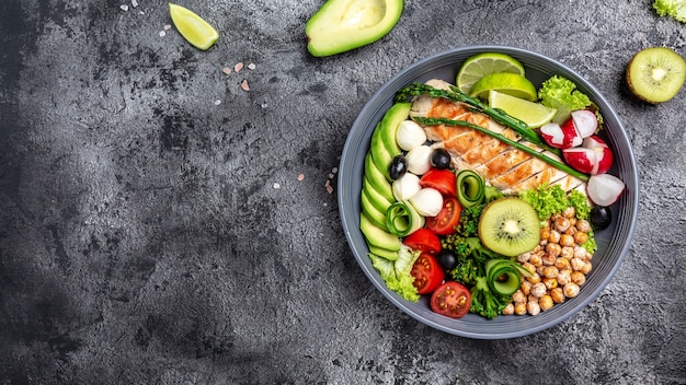 Pollo alla griglia, avocado, asparagi, ceci, broccoli, ravanello, pollo, cetriolo, pomodori, olive, mozzarella buddha bowl su sfondo scuro, vista dall'alto. delizioso concetto di cibo equilibrato