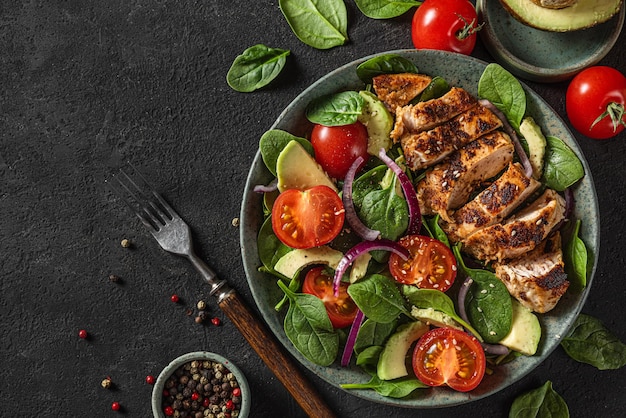 Grilled chicken breast fillet with spinach salad with avocado tomatoes and sesame seeds in a plate on dark background with fork top view with copy space