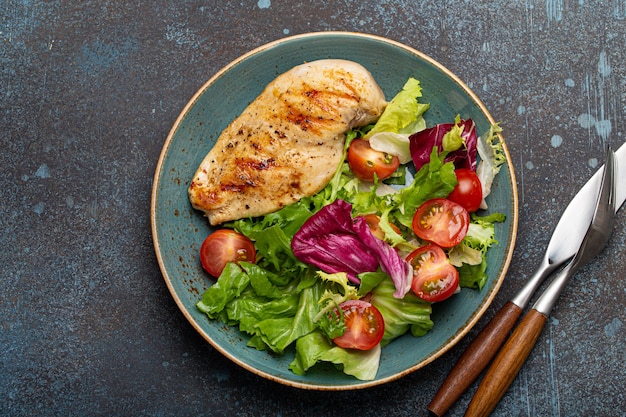 Grilled chicken breast fillet with vegetables salad of fresh tomatoes and salad leaves on blue plate on rustic stone background from above, healthy dinner or diet meal