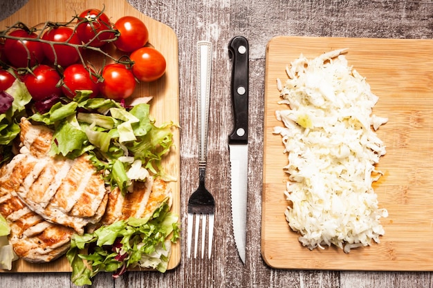 Petto di pollo alla griglia su insalata fresca tagliata accanto a un mazzetto di pomodori su una tavola di legno. sulla seconda tavola di legno c'è l'insalata di cavolo