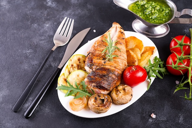 Grilled chicken breast on a plate with tomatoes, mushrooms and green sauce on a stone, flat lay