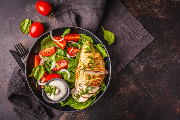 Grilled chicken breast salad with spinach, tomatoes and Caesar dressing, dark background.