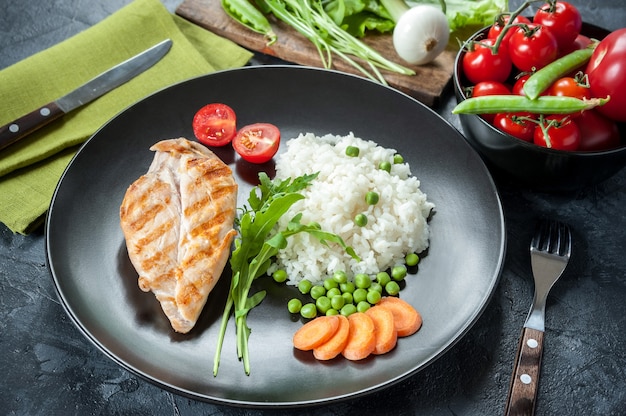 Grilled chicken fillet on plate with boiled rice. Raw vegetables, kitchen surface. Selective focus