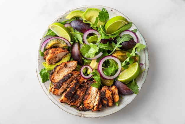 Grilled chicken fillet salad with arugula, avocado, plums, onion and lime in a plate on white background. Top view