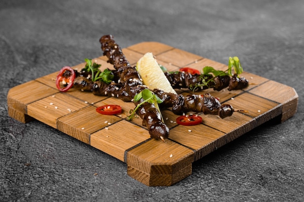 Grilled chicken hearts with lime on wooden skewer, garnished with chili, cilantro, sesame on gray surface.