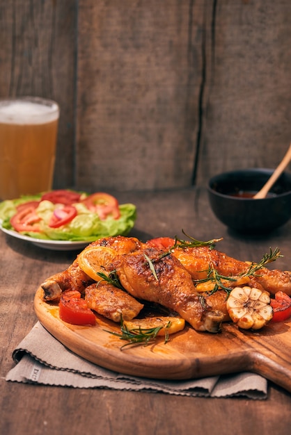 Grilled chicken legs roasted on the grill on wooden chopping board with tomato sauce in a bowl, fresh tomatoes and lettuce leaves, bitter pepper, glass mug of beer