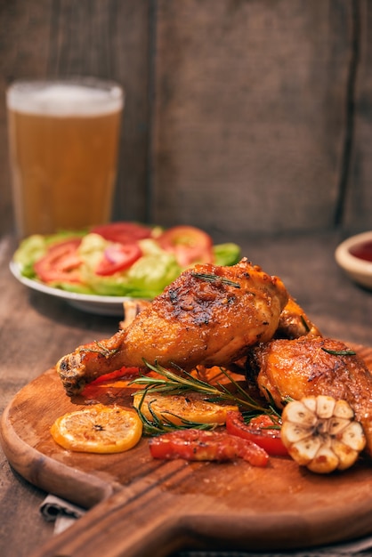 Grilled chicken legs roasted on the grill on wooden chopping board with tomato sauce in a bowl, fresh tomatoes and lettuce leaves, bitter pepper, glass mug of beer