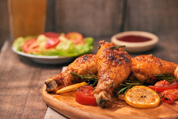 Grilled chicken legs roasted on the grill on wooden chopping board with tomato sauce in a bowl, fresh tomatoes and lettuce leaves, bitter pepper, glass mug of beer