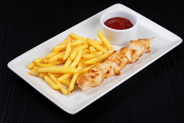 Grilled chicken shashlik meat with fried potatoes on white plate on black wooden background