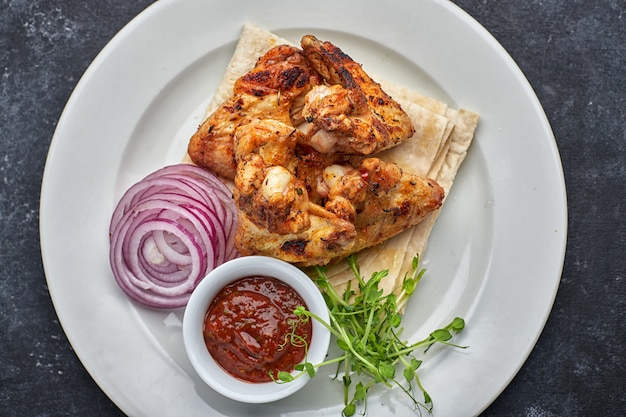 Grilled chicken wings with barbecue sauce, pita bread, microgreen and onion rings, on a white plate, against a dark table. Top view