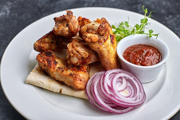 Grilled chicken wings with barbecue sauce, pita bread, microgreen and onion rings, on a white plate, against a dark table