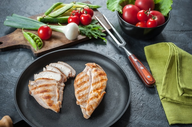 Grilled chiken fillet on frying pan. Raw vegetables in bowl, kitchen background. Selective focus