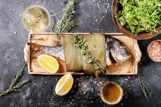 Foto pesce orata o orata eviscerato fresco cotto alla griglia servito con limone, insalata verde, rosmarino, olio d'oliva, vino bianco e sale rosa su un piatto di ceramica avvolto in foglie di bambù su una superficie scura.