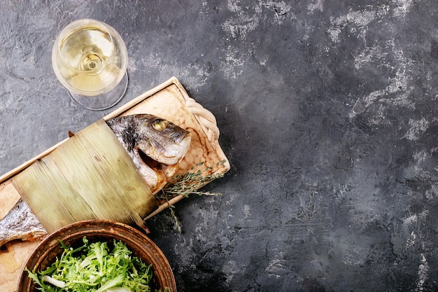 Pesce orata o orata eviscerato fresco cotto alla griglia servito con limone, insalata verde, rosmarino, olio d'oliva, vino bianco e sale rosa su un piatto di ceramica avvolto in foglie di bambù su una superficie scura.