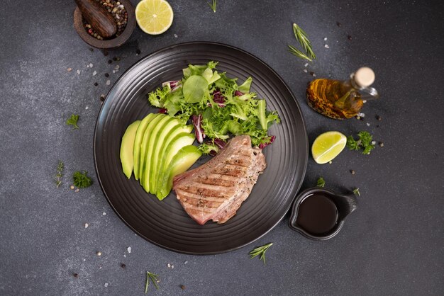 Grilled cooked piece of tuna fillet with sliced avocado and salad on black ceramic plate