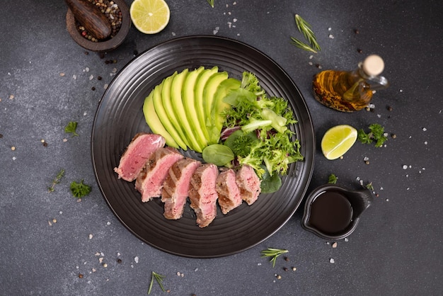 Grilled cooked piece of tuna fillet with sliced avocado and salad on black ceramic plate