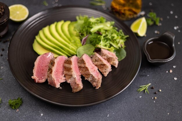 Grilled cooked piece of tuna fillet with sliced avocado and salad on black ceramic plate