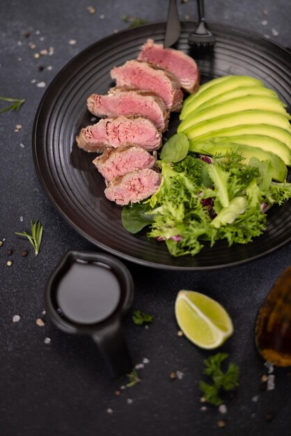 Grilled cooked piece of tuna fillet with sliced avocado and salad on black ceramic plate