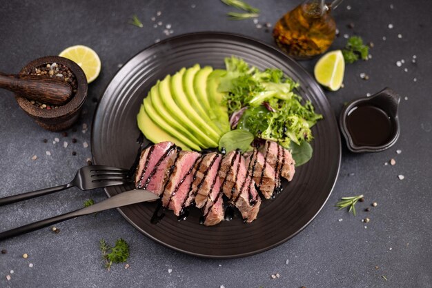 Grilled cooked piece of tuna fillet with sliced avocado and salad on black ceramic plate