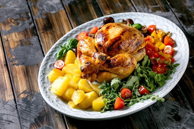 Grilled cooked whole chicken with vegetable garnish grilled bell pepper, onion, baked potatoes, cherry tomatoes, mushrooms and herbs in ceramic plate over dark wooden surface.