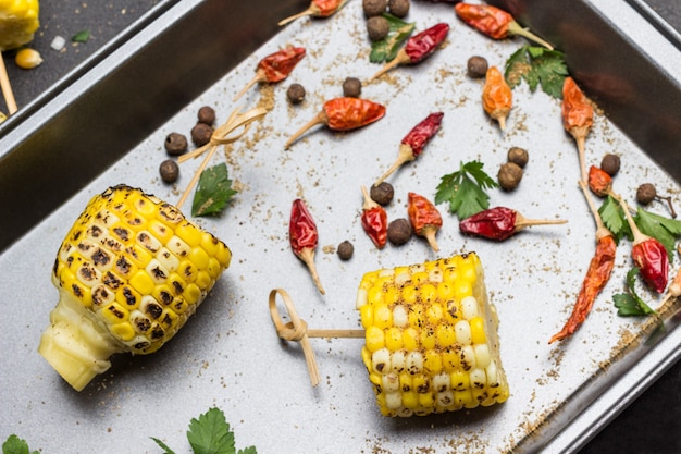 Grilled corn cobs on skewers. Dry red pepper pods and parsley leaves on tray. Black background. Flat lay