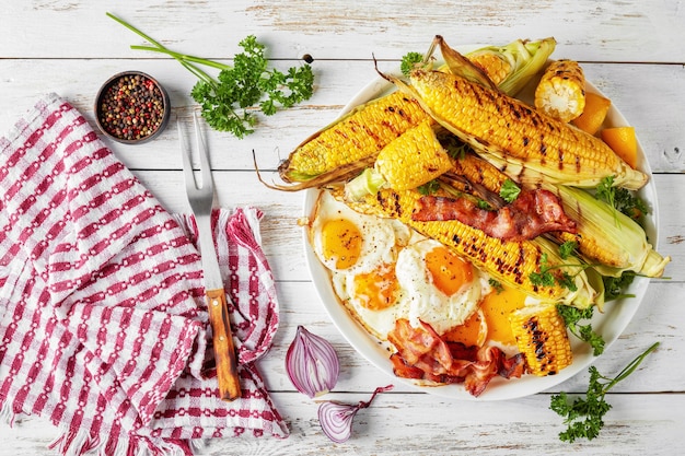 Grilled corn in cobs with fried eggs and bacon slices on a white dish on an old rustic table with napkin and fork, horizontal view from above, flat lay, close-up