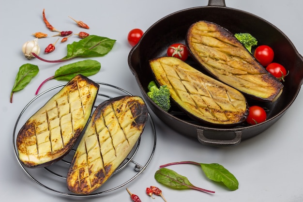 Grilled eggplants on wire rack and in frying pan. Chard leaves and garlic on the table.