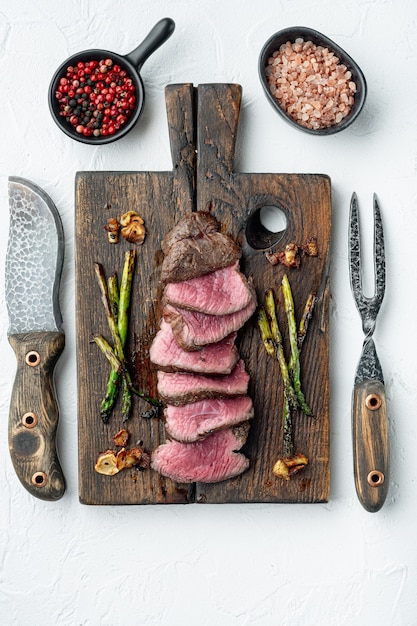 Grilled fillet beef steaks set, with onion and asparagus, on wooden serving board, with meat knife and fork, on white stone table, top view flat lay