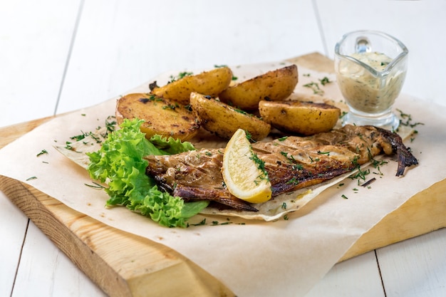 Филе рыбы на гриле с запеченным картофелем с соусом на деревянной доске. Белый деревянный фон