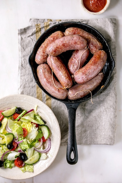 Photo grilled italian sausages salsiccia in cast-iron pan served with tomato sauce and plate of fresh vegetable salad. balanced dinner. flat lay