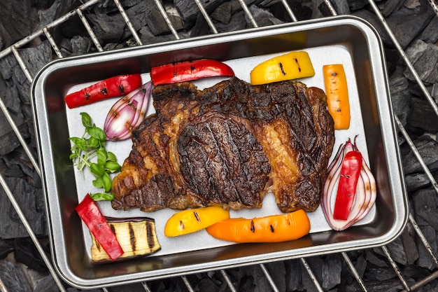 Grilled marbled beef steak on B-B-Q grill with vegetables on coals barbecue.