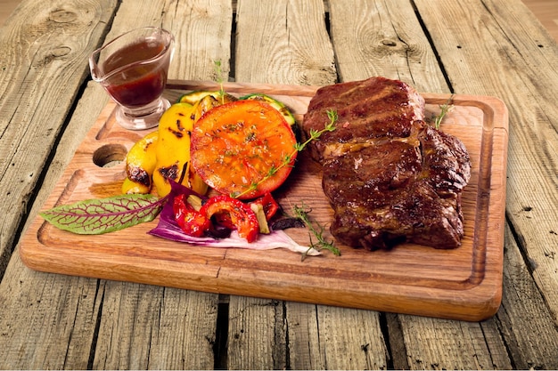 Grilled meat and vegetables with sauce and herbs on chopping board on wooden table background