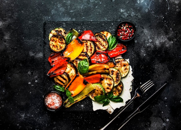 Grilled multicolored vegetables aubergines zucchini pepper with green basil on serving stone board on black background top view