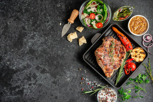 Grilled pork steak on cast iron pan with spices and vegetables on black. Top view