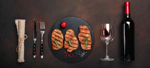 Grilled pork steaks on stone with bottle of wine, wine glass, knife and fork on rusty background. Top view with space for your text.