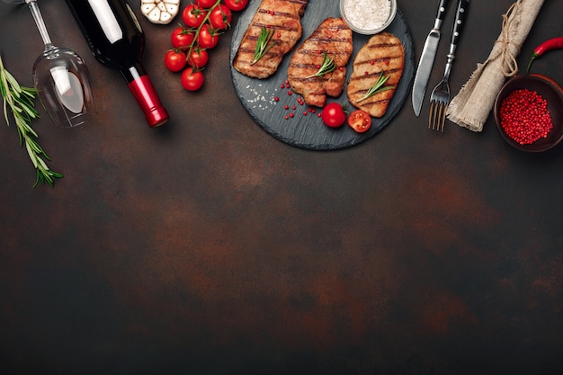 Photo grilled pork steaks on stone with bottle of wine, wine glass, knife and fork on rusty background