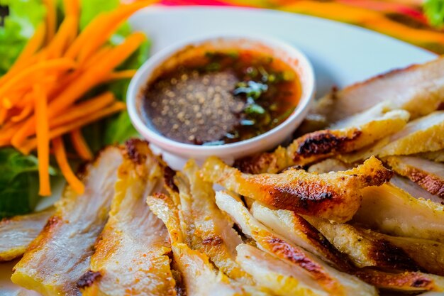 Grilled pork in a white dish with vegetables and dipping sauce on a wooden table.