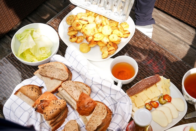 Grilled potatoes, salad, sliced bread, cup of tea, and cheese slices on plastic table outside - grilling concept.