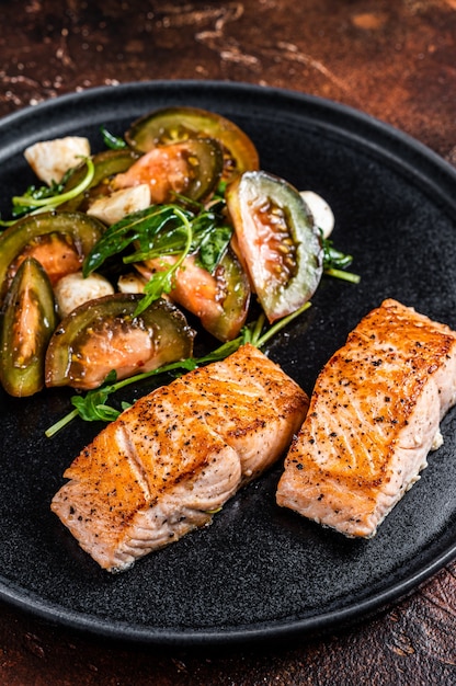 Photo grilled salmon fillet steaks with arugula and tomato salad on a plate. dark background. top view.