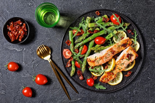 Grilled salmon fillets with lemon and caramelized bacon served with asparagus cherry tomatoes wild rocket salad on a black plate with golden cutlery, flat lay