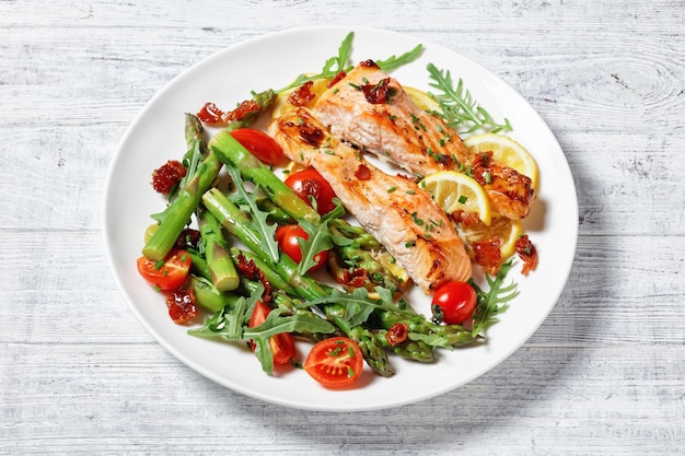 Grilled salmon fillets with lemon and caramelized bacon served with asparagus, cherry tomatoes, wild rocket salad on a white plate