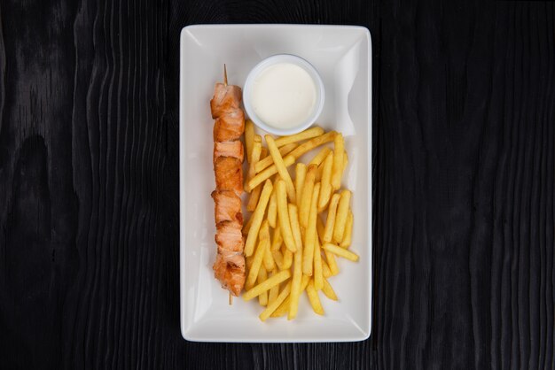 Grilled salmon fish shashlik with fried potatoes and sauce on white plate on black wooden background