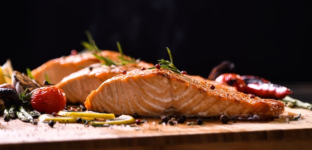 Grilled salmon fish and various vegetables on wooden table on black